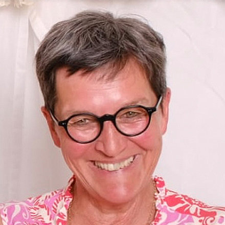 Portrait von Judith Bader-Reissing. Eine Frau mit kurzem, grauem Haar und rundem Brillengestell lächelt. Sie trägt ein Hemd mit rosa Muster. Der Hintergrund ist hell und einfarbig.