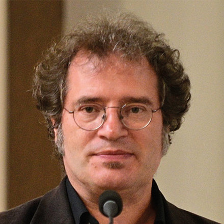 Ein Porträtfoto eines Mannes mit lockigem braunem Haar, Brille und einem kleinen Ziegenbart. Er trägt ein dunkles Hemd und eine dunkle Jacke und blickt direkt in die Kamera. Vor ihm ist ein Mikrofon zu sehen.