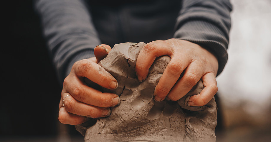 Nahaufnahme von zwei Händen, die einen großen Klumpen braungrauen Tons kneten oder formen. Die Hände sind mit Ton bedeckt, und an einem Finger ist ein Ring zu sehen. Die Person trägt einen grauen Langarmpullover.