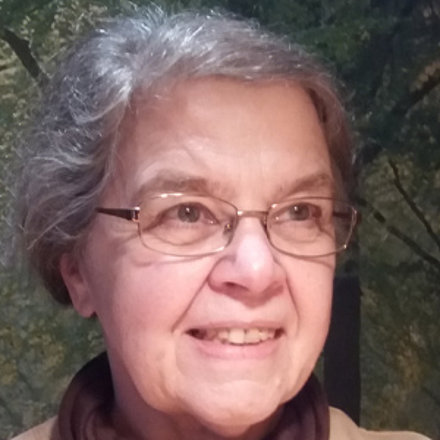 Eine ältere Frau mit grauen, lockigen Haaren und einer Brille lächelt in die Kamera. Sie trägt einen braunen Rollkragenpullover oder Schal über einem hellen Oberteil. Der Hintergrund ist unscharf und zeigt einige Bäume und Büsche.