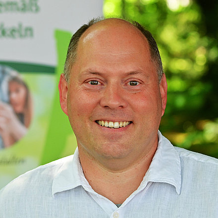 Portrait von Stefan Weiland, ein lächelnder, glatzköpfigen Mannes mit heller Haut, der ein weißes Hemd trägt. Im Hintergrund sind unscharfe grüne Pflanzen und ein Banner mit teilweise sichtbarem Text.