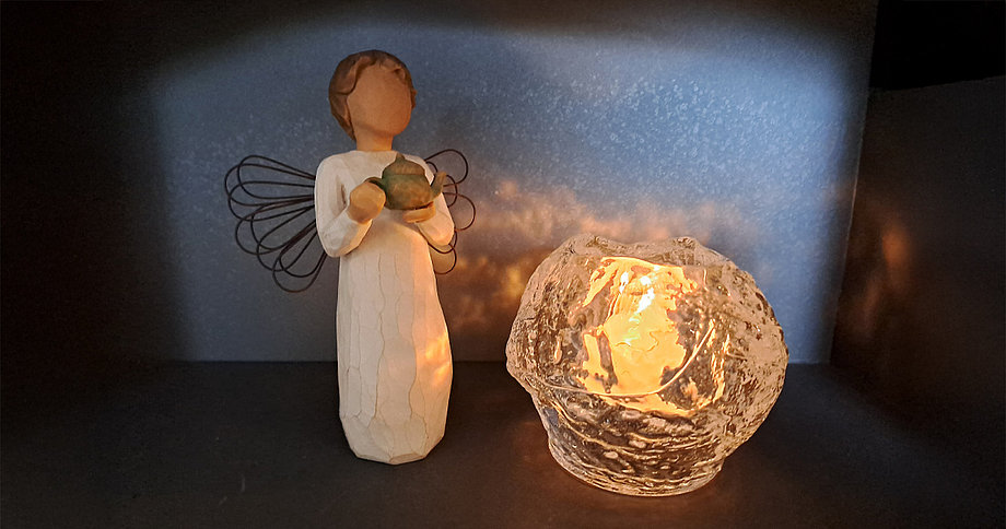 Eine kleine, holzgeschnitzte Engelsfigur mit Drahtflügeln, die einen grünen Kelch hält, steht neben einem leuchtenden Teelicht in einem strukturierten Glaswindlicht. Beide Objekte sind auf einer dunklen Oberfläche vor einem dunklen Hintergrund platziert, wobei das Licht des Teelichts die Engelsfigur von vorne beleuchtet.