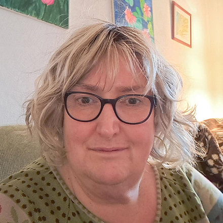 Porträt einer Frau mittleren Alters mit blondem, welligem Haar und schwarzer Brille, die leicht lächelt und direkt in die Kamera blickt. Sie trägt ein grünes Oberteil mit einem Muster. Im Hintergrund sind eine Couch und zwei gerahmte Bilder an einer hellen Wand zu sehen.