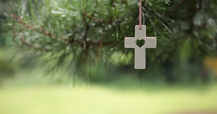 Ein weißes Kreuz mit einem herzförmigen Ausschnitt in der Mitte hängt an einem dünnen Band vor unscharfem Hintergrund aus grünen Tannenzweigen und einer hellgrünen Wiese.